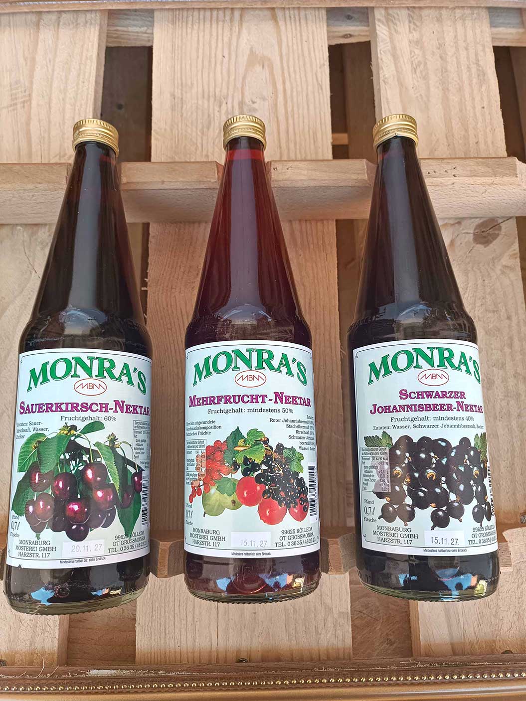 Drei Flaschen Monra's Saft auf einem Holztisch, alle mit Fruchtetiketten.